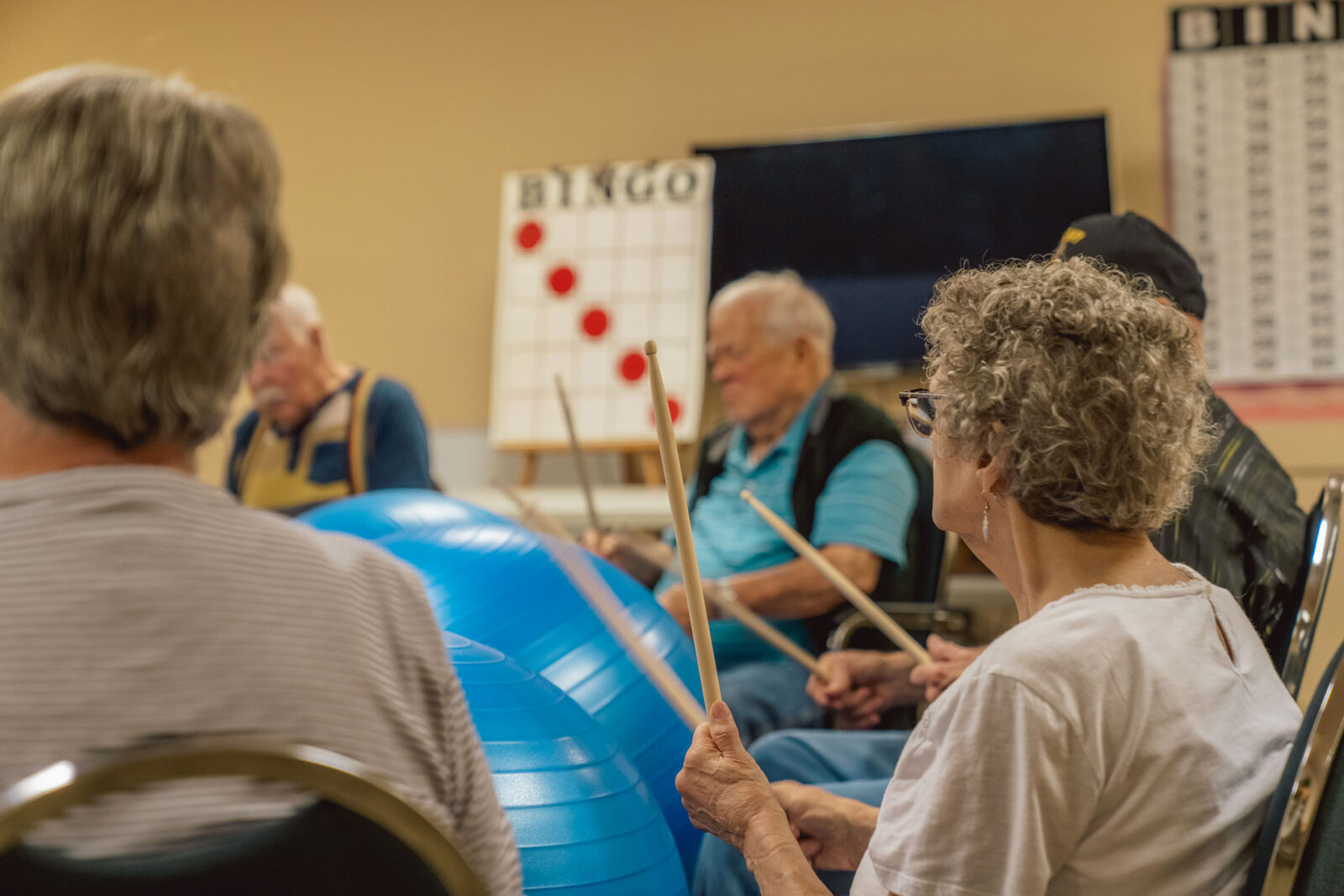Seniors participating in a rhythmic activity, holding sticks near a Bingo board.