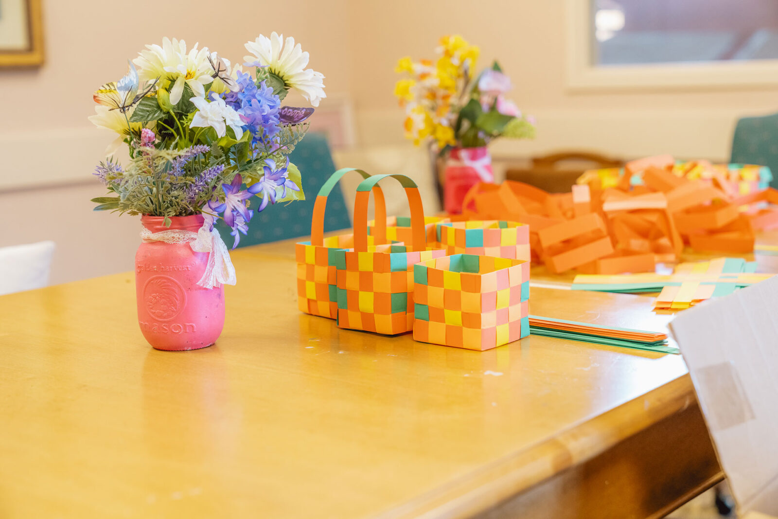 A colorful craft table with paper baskets and a flower arrangement in a pink jar.