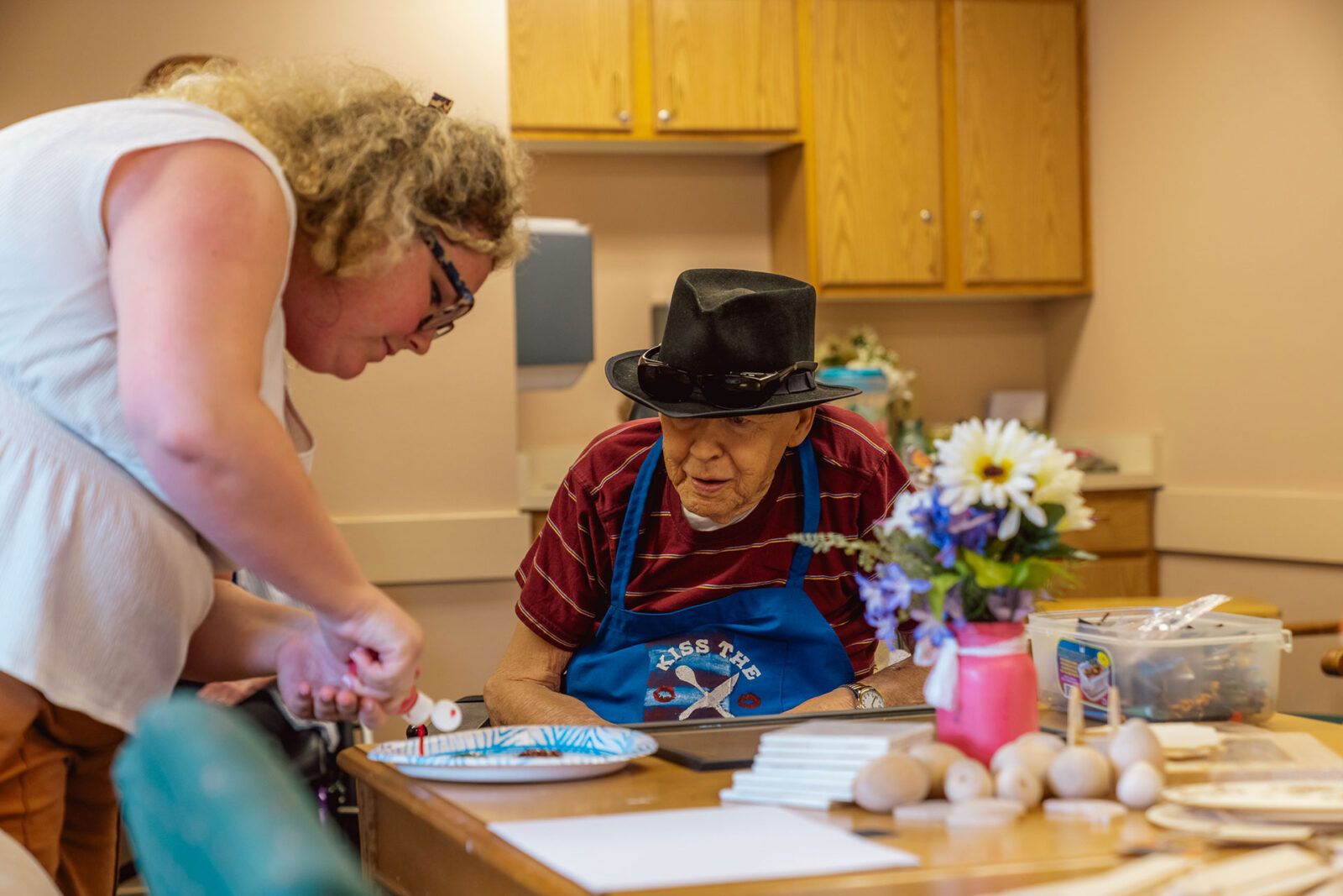 A woman helps an elderly man with a craft activity at a table.