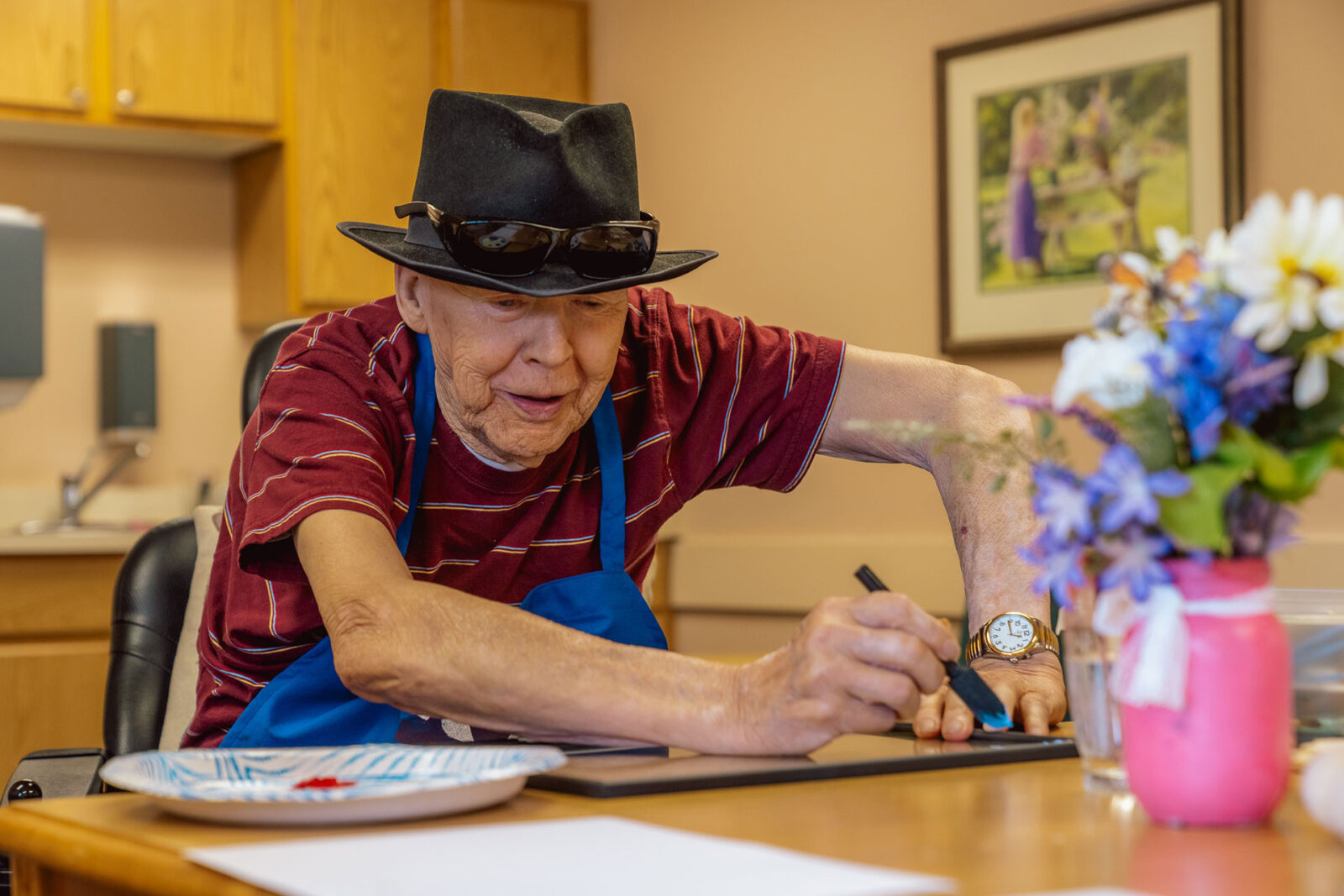 An elderly man in a hat sketches art, surrounded by flowers and a colorful workspace.